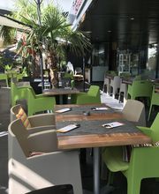 Außenterrasse eines Cafés mit grünen Stühlen, Holztischen und Palmen in Töpfen.