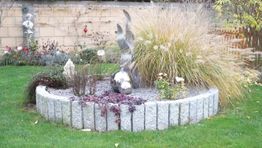 Rundbeet mit Ziergräsern und Steinstatuen im Garten, umgeben von Rasen und Mauer im Hintergrund.