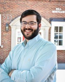 Smiling person in glasses and checked shirt poses confidently in front of a brick building.