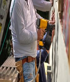 Workers using a nail gun on a construction site wall, wearing tool belts and white shirts.