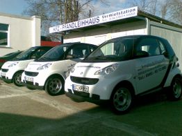 Drei weiße Smart-Autos mit Vermietstation im Hintergrund, beschriftet mit Telefonnummer.