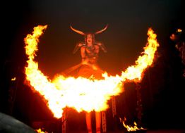 Person mit Hörnern hinter brennendem Halbkreis in der Dunkelheit, dramatische Inszenierung.