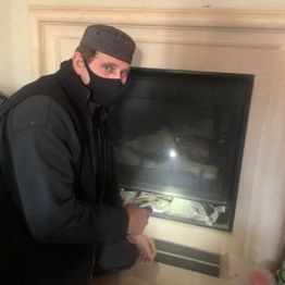 Person in a mask and cap kneeling by a fireplace with a flashlight inside.