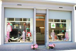 Fassade eines Ladens mit Schaufenstern, Kinderkleidung und Spielzeug; Blumendeko am Eingang.