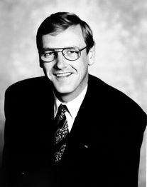 Ein Mann mit Brille im Anzug, lächelt vor einem neutralen Hintergrund. Schwarz-weiß Foto.