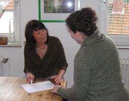 Zwei Frauen reden miteinander an einem Tisch. Eine Frau zeigt auf ein Dokument.