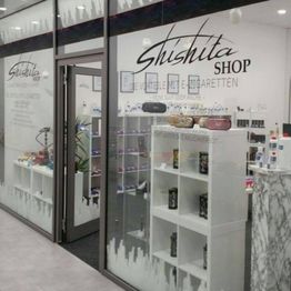Schaufenster eines E-Zigarettenladens mit Regalen voller Produkte und Pflanzen im Inneren.