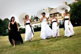 Bridesmaids in black and white dresses walk on grass, holding white parasols near a house.