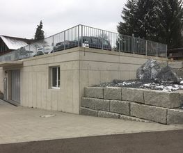 Haus mit Betongarage und steiniger Landschaft, oben sind geparkte Autos sichtbar.