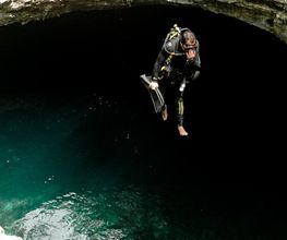 Persona buceando en cenote, colgada sobre el agua azul, rodeada de paredes rocosas.