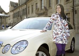 Frau steht in bunter Jacke neben einem weißen Auto auf einer städtischen Straße.