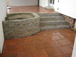 Steintreppe neben rundem Brunnen auf Terrasse mit roten Fliesen.