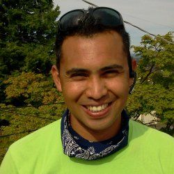 Smiling person outdoors wearing a green shirt, bandana, and sunglasses on their head.