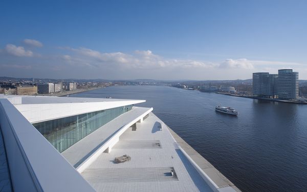 Ein modernes Unternehmensgebäude an einem breiten Fluss. In der Nähe fährt ein Boot vorbei. Im Hintergrund ist die Skyline einer Großstadt zu erkennen.