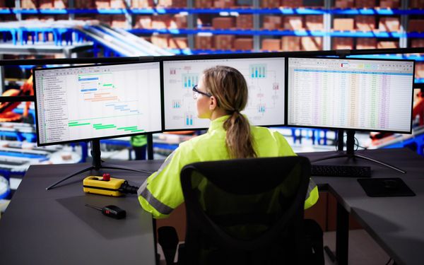 Eine Spediteurin sitz vor drei Computer Monitoren und steuert die logistischen Prozesse des Unternehmens.