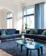 Modernes Wohnzimmer mit blauen Sofas, Vorhängen und Teppich. Helle Fenster, Kunstwerke an der Wand.