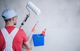 Painter with a roller and bucket stands by a blank wall, wearing a cap and red shirt.