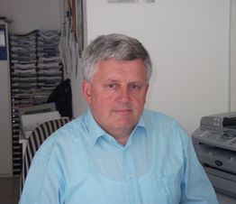 Ein Mann im blauen Hemd sitzt in einem Büro, mit Ordnern und Drucker im Hintergrund.