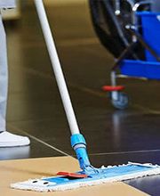 Person wischt Boden, mit einem blauen Wischmopp und Eimer auf Rollen im Hintergrund.