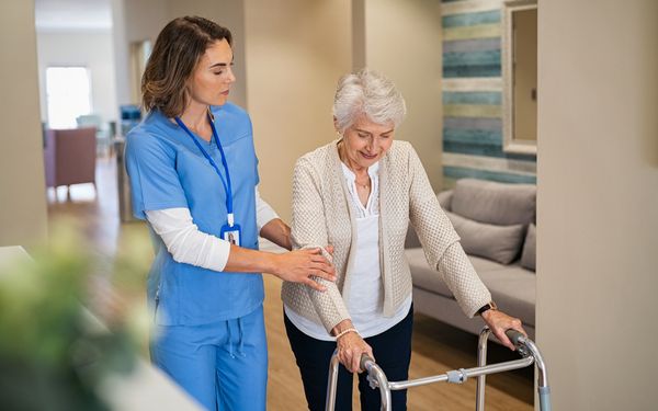 Eine Pflegefachassistentin stützt einer Seniorin mit Rollator in einem Pflegeheim.