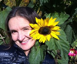 Eine Frau lächelt neben einer großen, gelben Sonnenblume im Freien.