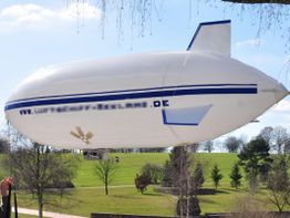 Ein weißes Luftschiff mit blauen Streifen fliegt über einen grünen Park unter blauem Himmel.