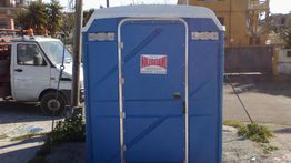 Bagno chimico blu all'aperto con un cartello di noleggio. Furgone bianco parcheggiato a sinistra.