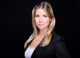 Eine Frau mit blonden Haaren in schwarzem Blazer schaut ernst in die Kamera vor schwarzem Hintergrund.