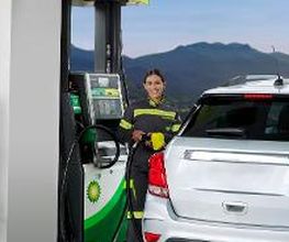 Mujer en uniforme suministra gasolina a un auto blanco en una estación de servicio.