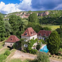 Maison en pierre avec piscine, entourée de verdure, montagnes en arrière-plan.