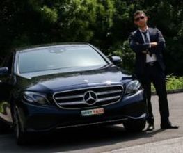 Ein Mann in Anzug steht neben einem schwarzen Mercedes auf einer Straße, hinter ihm Bäume.