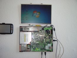 Offener Laptop an der Wand montiert, zeigt ein Motherboard und einen Bildschirm mit Windows-Logo.