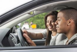 Two people smiling in a car, one at the wheel, suggesting a driving lesson or road trip.