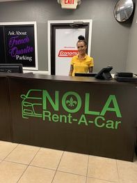 Woman at NOLA Rent-A-Car desk, yellow shirt, sign about French Quarter parking above.