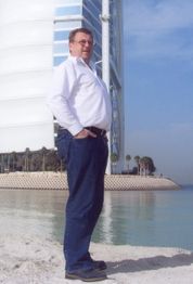 Mann in weißem Hemd steht am Wasser vor modernem Gebäude und Palmen unter blauem Himmel.