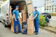 Two movers in blue uniforms unload cardboard boxes from a van using a dolly.