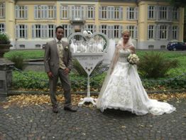 Brautpaar vor einem eleganten Gebäude mit Herzaufsteller und "Viel Glück"-Schild.