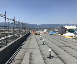 Baustelle auf einem Flachdach mit Gerüsten, Bergen im Hintergrund und blauem Himmel.
