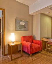 Sala de hotel com poltrona vermelha, mesa, cadeiras e vista parcial do quarto com cama branca.