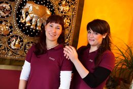 Zwei Frauen in burgunder T-Shirts lächeln vor einem kunstvollen Wandteppich mit Elefantendesign.