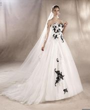 Braut in weißem Kleid mit schwarzen Blumenmustern und langem Schleier vor neutralem Hintergrund.