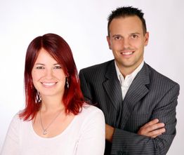 Zwei lächelnde Menschen, eine Frau mit roten Haaren und ein Mann im Anzug, vor weißem Hintergrund.