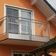 Balkon mit Metallgeländer an orangefarbenem Haus, darüber Fenster mit weißen Vorhängen.