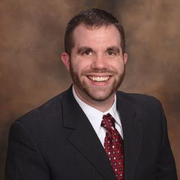 Smiling man in a suit with a red tie, against a brown background.