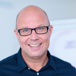 Ein lächelnder Mann mit Brille und Glatze in einem dunklen Hemd vor verschwommenem Hintergrund.