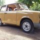 Beiger Trabant-Pkw parkt auf einer Einfahrt, umgeben von Bäumen und einem Haus im Hintergrund.
