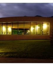 Rundes Gebäude mit großen Fenstern, nachts beleuchtet, vor einem bewölkten Himmel.