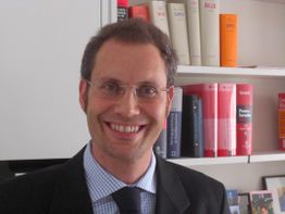 Eine Person mit Brille lächelt vor einem Bücherregal.