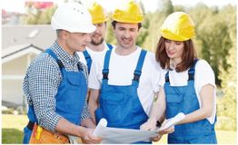 Construction workers in blue overalls and hard hats examine a blueprint outdoors.