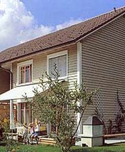 Zweistöckiges Haus mit Garten, Sonnenschutz und Holzwandverkleidung, blauer Himmel im Hintergrund.